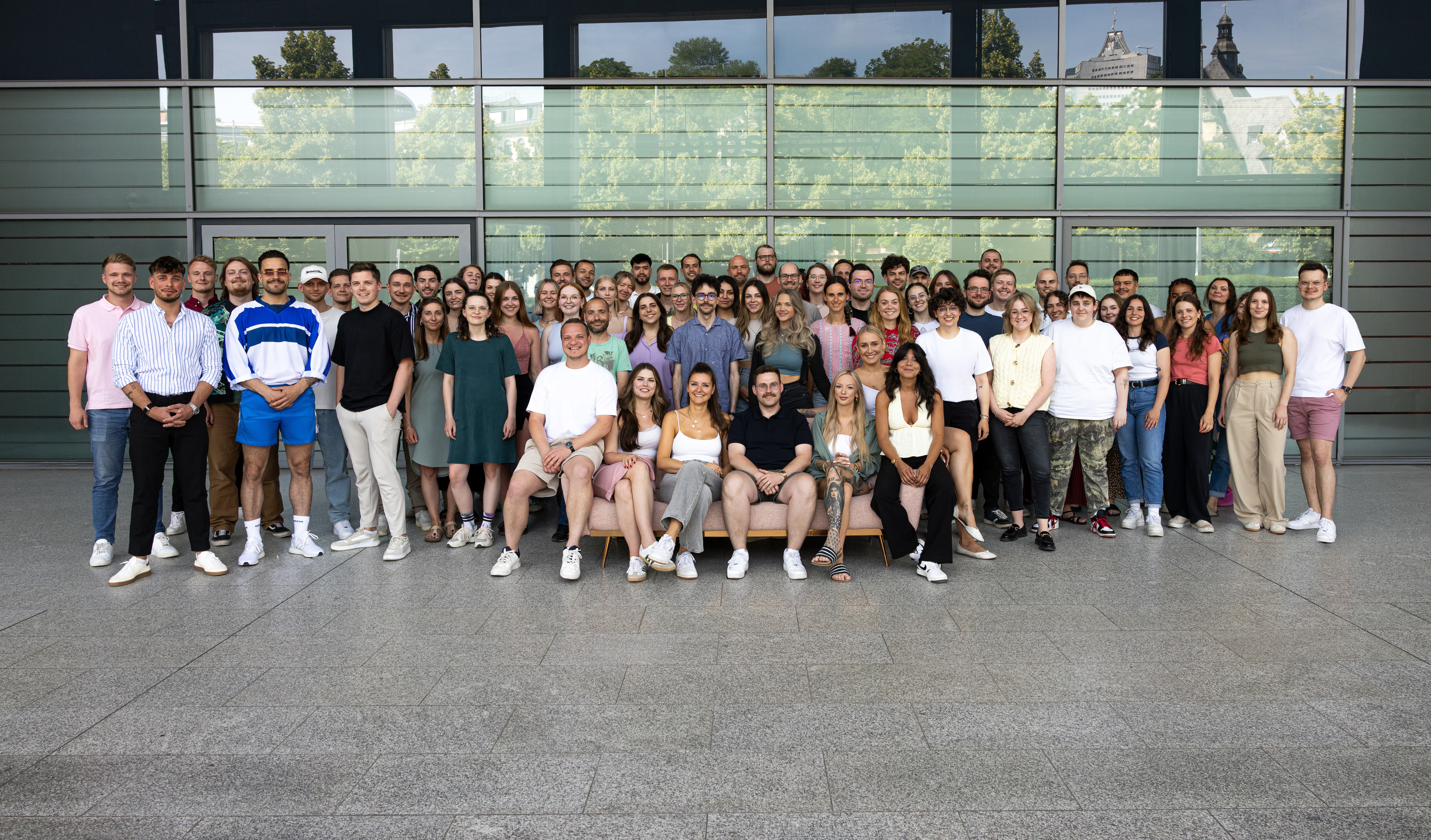 Große Gruppe von etwa 70 Personen, die in mehreren Reihen vor einem modernen Glasgebäude stehen und sitzen, lächelnd und in lässiger Kleidung.